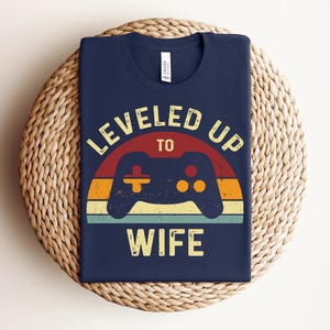 May include: Navy blue t-shirt with a retro-style graphic of a video game controller and the text "LEVELED UP TO WIFE".
