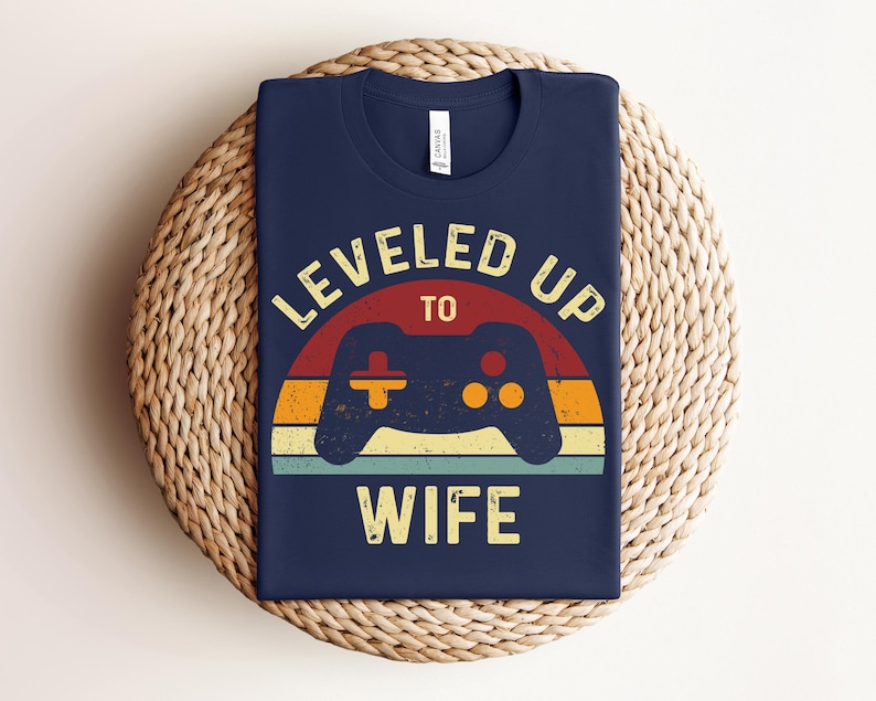 May include: Navy blue t-shirt with a retro-style graphic of a video game controller and the text "LEVELED UP TO WIFE".