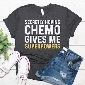 May include: A dark gray t-shirt with the text "Secretly Hoping Chemo Gives Me Superpowers" in white and yellow lettering. The shirt is laid flat on a wooden surface with a pair of white sneakers and denim shorts.