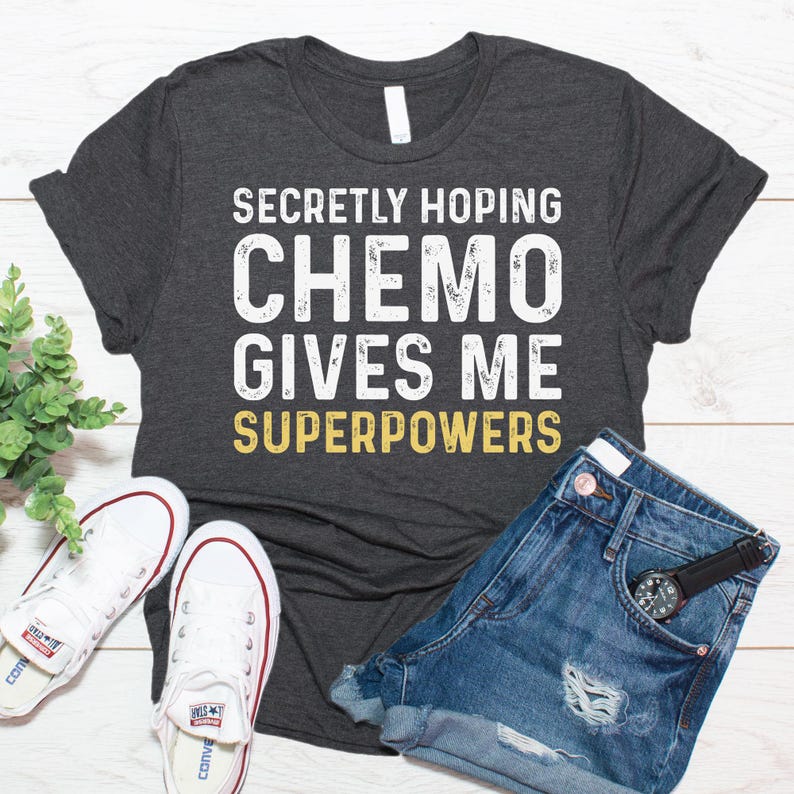 May include: A dark gray t-shirt with the text "Secretly Hoping Chemo Gives Me Superpowers" in white and yellow lettering. The shirt is laid flat on a wooden surface with a pair of white sneakers and denim shorts.