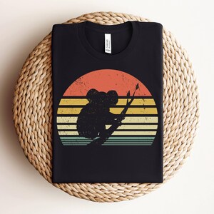 May include: Black t-shirt with a vintage-style graphic of a koala sitting on a branch in front of a sunset. The graphic has a retro color palette with shades of orange, yellow, and blue.