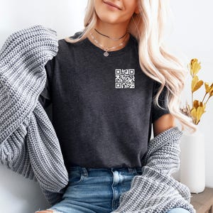 May include: A dark charcoal grey short-sleeved t-shirt featuring a small white QR code graphic printed on the upper left chest area. The shirt is worn with a chunky grey knit cardigan and blue denim jeans, creating a layered casual look.