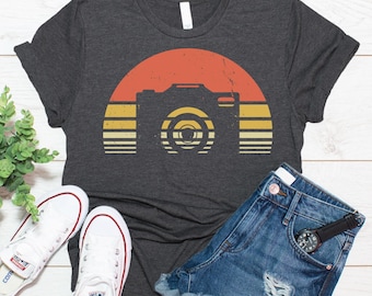 May include: A dark charcoal gray t-shirt featuring a graphic of a camera centered within a distressed sunset design composed of horizontal stripes in shades of orange, yellow, and cream. The shirt is paired with blue denim shorts and white sneakers.