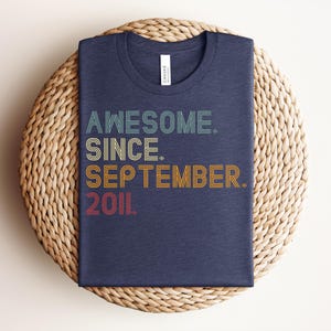 May include: A dark blue crew neck t-shirt featuring the text "AWESOME. SINCE. SEPTEMBER. 2011." in a multi-colored, retro-inspired linear font. The words are stacked vertically, with each line in a different color including teal, yellow, orange, and red.