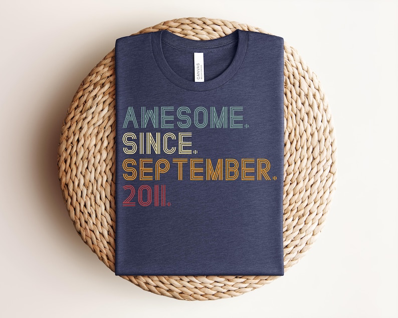 May include: A dark blue crew neck t-shirt featuring the text "AWESOME. SINCE. SEPTEMBER. 2011." in a multi-colored, retro-inspired linear font. The words are stacked vertically, with each line in a different color including teal, yellow, orange, and red.