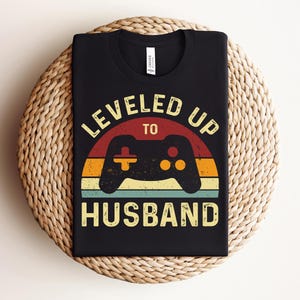 May include: Black t-shirt featuring a retro-style graphic that reads "Leveled Up To Husband" with a video game controller in the centre.