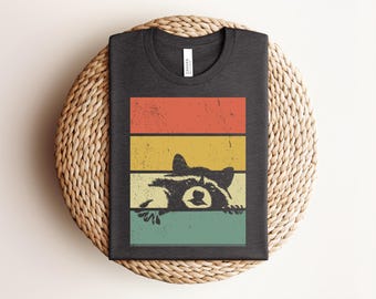Puede incluir: Una camiseta gris oscuro con un estampado gráfico de un mapache asomándose sobre una barra horizontal. El diseño incluye cuatro franjas rectangulares apiladas en naranja, amarillo y verde azulado con una textura desgastada y vintage.