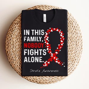 Może przedstawiać: Czarna koszulka z białym napisem "IN THIS FAMILY, NOBODY FIGHTS ALONE." oraz słowem "NOBODY" w kolorze czerwonym. Po prawej stronie znajduje się czerwona wstążka świadomości z małych białych i czerwonych serc oraz napis "Stroke Awareness".