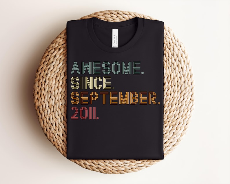 May include: A black crew neck t-shirt featuring the text AWESOME. SINCE. SEPTEMBER. 2011. printed in a multi-line, retro-inspired font. The lettering uses a color palette of teal, yellow, orange, and red, with each line having a distinct color.