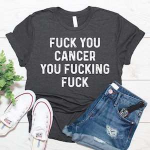 May include: A dark charcoal grey short-sleeve t-shirt featuring the white distressed-style text "FUCK YOU CANCER YOU FUCKING FUCK" printed across the chest. The shirt is displayed alongside a pair of blue denim shorts and white trainers.
