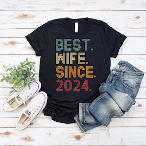 Op de afbeelding: Een zwart T-shirt met korte mouwen met de tekst BEST. WIFE. SINCE. 2024. in een meerkleurig, gestreept lettertype in retrostijl. Het T-shirt wordt getoond naast een blauwe spijkerbroek en witte canvas sneakers met witte veters.