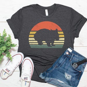 May include: A dark charcoal gray short-sleeve t-shirt featuring a graphic of a black wombat silhouette centered over a distressed, multi-colored sunset design with horizontal stripes in shades of orange, yellow, and muted green.