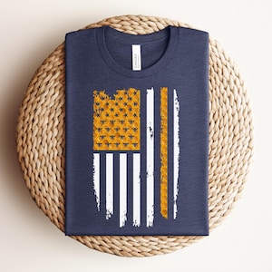 May include: A navy blue t-shirt featuring a graphic design of an American flag. The flag's canton and one stripe are composed of a yellow honeycomb pattern with small bee silhouettes, while the remaining stripes are rendered in white.