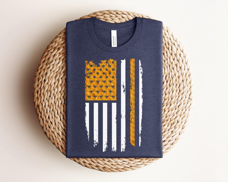 May include: A navy blue t-shirt featuring a graphic design of an American flag. The flag's canton and one stripe are composed of a yellow honeycomb pattern with small bee silhouettes, while the remaining stripes are rendered in white.