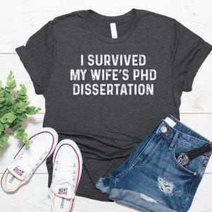 May include: A dark charcoal gray short-sleeve t-shirt featuring the white distressed text "I SURVIVED MY WIFE'S PHD DISSERTATION" printed across the chest. The shirt is displayed alongside white canvas sneakers, blue denim shorts, and a small plant.