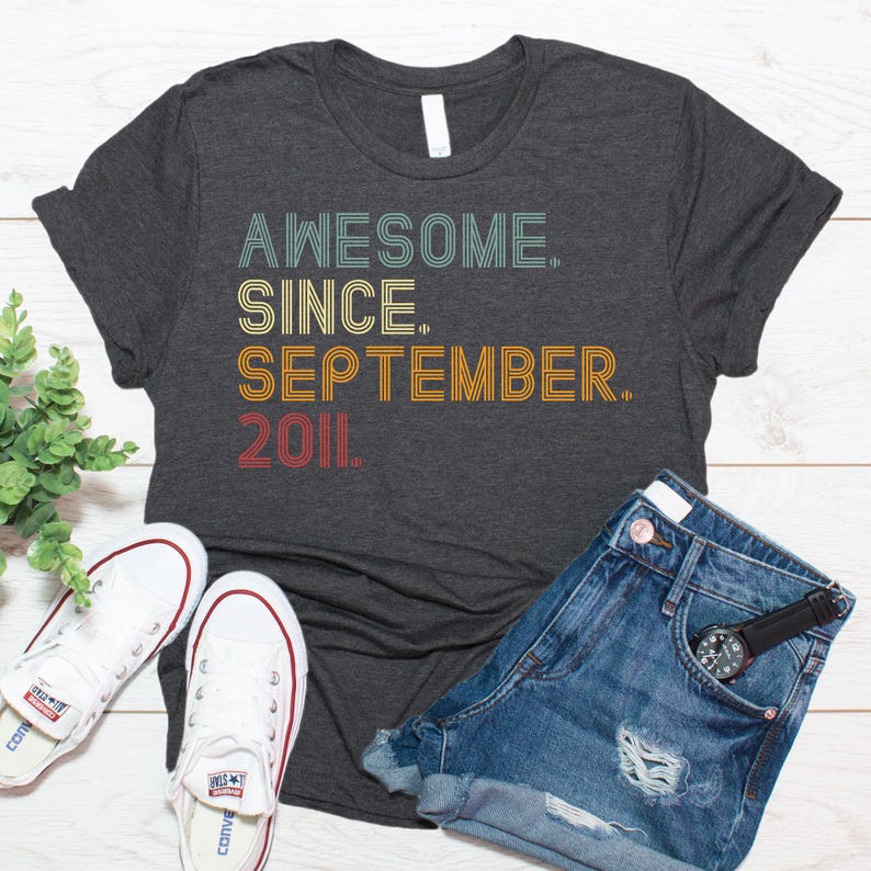 May include: A dark heather gray short-sleeve t-shirt featuring the text "AWESOME. SINCE. SEPTEMBER. 2011." in a retro-style, multi-colored, layered font. The shirt is paired with blue denim shorts and white canvas sneakers with red and blue stripes.