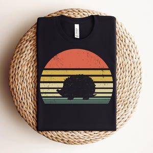 K&ouml;nnte beinhalten: Schwarzes T-Shirt mit einem Retro-Sonnenuntergangsdesign mit einer Silhouette eines Igels. Der Sonnenuntergang hat orange, gelbe und blaue Streifen.