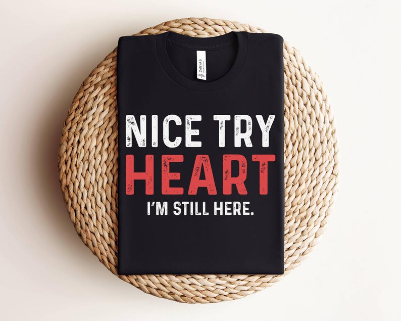 Puede incluir: Camiseta negra con texto blanco que dice "Nice Try Heart I'm Still Here."