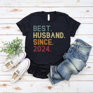 Op de afbeelding: Een zwart T-shirt met korte mouwen met de tekst "BEST. HUSBAND. SINCE. 2024." in een meerkleurig, retro-geïnspireerd lettertype. Het T-shirt wordt getoond naast een blauwe spijkerbroek en witte canvas sneakers met witte veters.