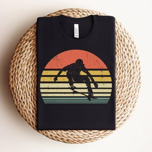 May include: A black t-shirt with a retro-style graphic of a person skateboarding in front of a sunset. The graphic has a faded, vintage look with a yellow, orange, and red sunset.