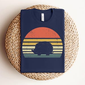K&ouml;nnte beinhalten: Ein marineblaues T-Shirt mit einem Retro-Sonnenuntergangsdesign, das eine Silhouette eines Igels zeigt. Der Sonnenuntergang ist in Orange-, Gelb- und Gr&uuml;nt&ouml;nen gehalten.