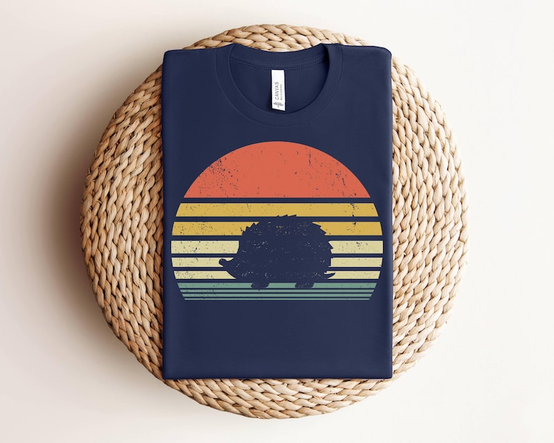 K&ouml;nnte beinhalten: Ein marineblaues T-Shirt mit einem Retro-Sonnenuntergangsdesign, das eine Silhouette eines Igels zeigt. Der Sonnenuntergang ist in Orange-, Gelb- und Gr&uuml;nt&ouml;nen gehalten.