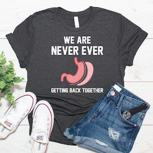 May include: A dark heather gray short-sleeve t-shirt featuring the white text "WE ARE NEVER EVER GETTING BACK TOGETHER" and a graphic illustration of a human stomach with a dashed line indicating a surgical procedure in the center.