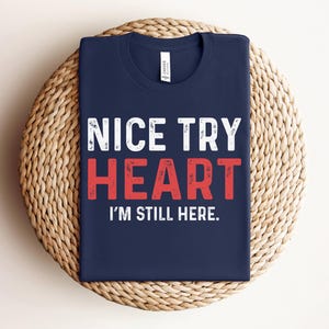 Puede incluir: Una camiseta azul marino con texto blanco que dice "NICE TRY HEART I'M STILL HERE."