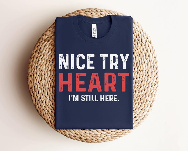 Puede incluir: Una camiseta azul marino con texto blanco que dice "NICE TRY HEART I'M STILL HERE."