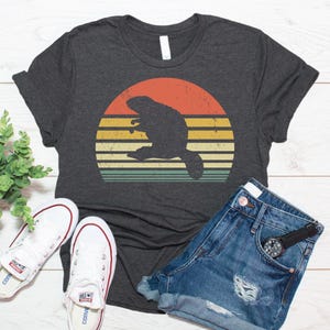 Könnte beinhalten: Ein dunkelgrau meliertes Kurzarm-T-Shirt mit einer Grafik der Silhouette eines Bibers, zentriert über einem abgenutzten, mehrfarbigen Sonnenuntergangskreis mit horizontalen Streifen in Orange, Gelb und Petrol.