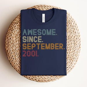 May include: A navy blue t-shirt featuring the text AWESOME. SINCE. SEPTEMBER. 2001. printed in a retro, multi-colored, line-art style font. The shirt has a crew neckline and a white brand label visible at the inner collar area.
