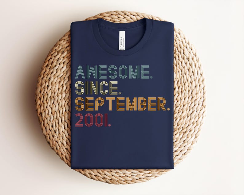 May include: A navy blue t-shirt featuring the text AWESOME. SINCE. SEPTEMBER. 2001. printed in a retro, multi-colored, line-art style font. The shirt has a crew neckline and a white brand label visible at the inner collar area.