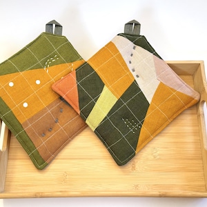 May include: Two square, quilted pot holders with geometric patterns in shades of green, orange, and brown. Each has a loop for hanging. They rest on a light brown wooden serving tray.