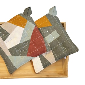 Puede incluir: Dos agarraderas cuadradas y acolchadas con diseños geométricos de patchwork en tonos verdes, naranjas, rojos y beige. Se encuentran sobre una bandeja de madera marrón claro con asas. Las agarraderas tienen lazos para colgar.