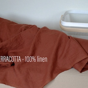 Peut inclure: Un rouleau de tissu en lin 100% de couleur terre cuite. Le tissu est plié et roulé, avec l'extrémité du rouleau visible. Le tissu est posé sur une surface brun clair.