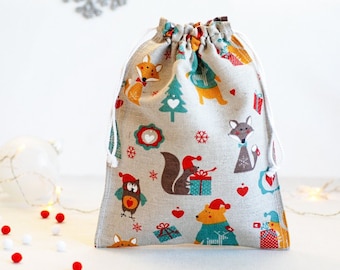 May include: A beige drawstring bag with a Christmas theme. The bag is decorated with a pattern of foxes, owls, squirrels, and presents. The pattern is in shades of red, green, blue, and yellow.
