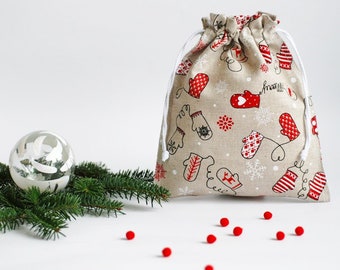May include: A beige drawstring bag with a white cord. The bag is decorated with a pattern of red and white mittens, snowflakes, and hearts. The bag is sitting on a white surface with a few red beads scattered around it.
