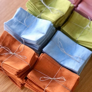 May include: Stacks of folded linen napkins in various colours, including light blue, green, orange, and pink. Each stack is tied with twine.