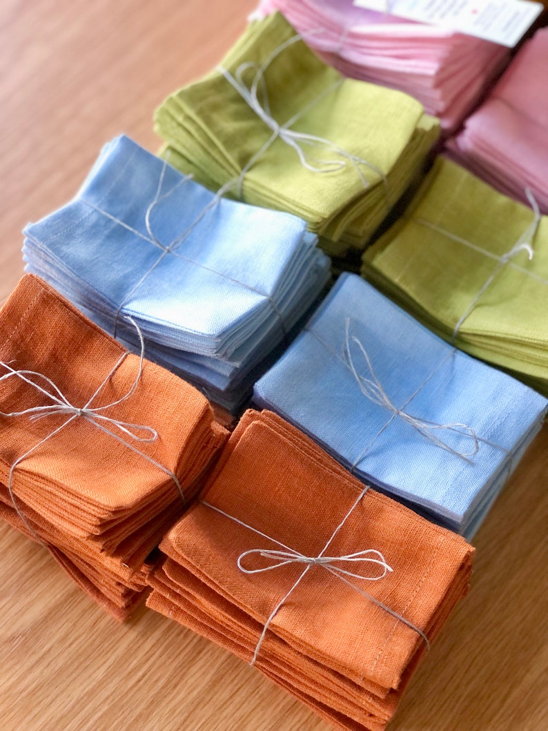 May include: Stacks of folded linen napkins in various colours, including light blue, green, orange, and pink. Each stack is tied with twine.