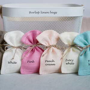May include: Six small burlap linen bags in different colors, including grey, white, pink, peach cream, ivory white, and teal blue. The bags are arranged in a row and labeled with their respective colors.
