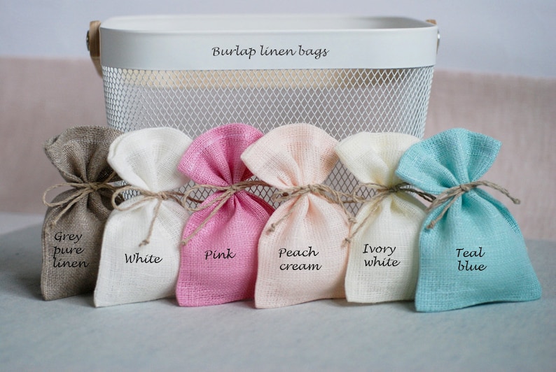May include: Six small burlap linen bags in different colors, including grey, white, pink, peach cream, ivory white, and teal blue. The bags are arranged in a row and labeled with their respective colors.