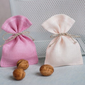 May include: Two small fabric gift bags, one pink and one beige, tied with twine. The bags are sitting on a light gray surface with three walnuts beside them.