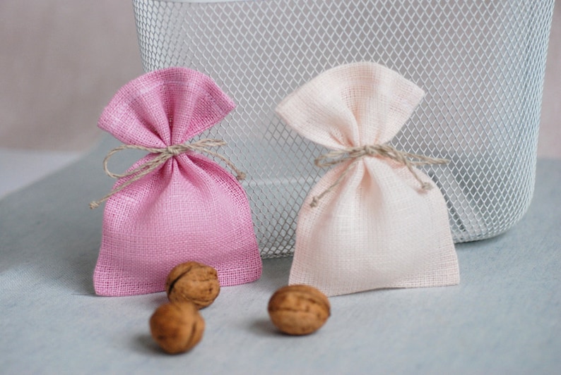 May include: Two small fabric gift bags, one pink and one beige, tied with twine. The bags are sitting on a light gray surface with three walnuts beside them.
