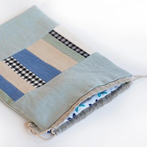 Può includere: Una borsa a cordoncino blu chiaro realizzata in tessuto patchwork con un motivo a quadri. La borsa è piegata e il cordoncino è visibile.