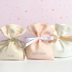 May include: Three small fabric gift bags in cream, light peach, and ivory colours. The bags are tied with different coloured ribbons and have the colour names printed on the front.