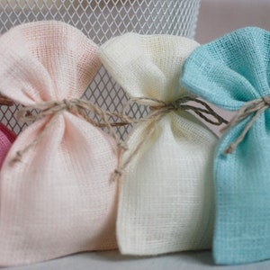 May include: Four small fabric gift bags in pink, cream, white, and light blue. The bags are tied with twine and are made of a soft, textured fabric.