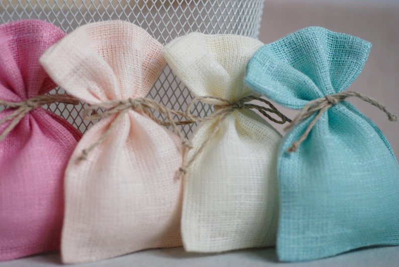 May include: Four small fabric gift bags in pink, cream, white, and light blue. The bags are tied with twine and are made of a soft, textured fabric.