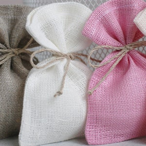 May include: Three small burlap gift bags tied with twine. The bags are beige, white, and pink.