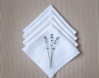 May include: Four white linen napkins folded in a square shape with a sprig of lavender flowers on top.