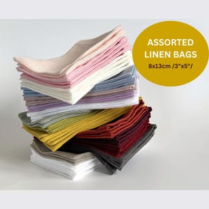 Könnte beinhalten: Ein Stapel verschiedener Leinentaschen in verschiedenen Farben, darunter Rosa, Weiß, Blau und Gelb. Die Taschen sind ordentlich gefaltet und gestapelt, mit dem Text "ASSORTED LINEN BAGS" und den Maßen 8x13cm im Bild.